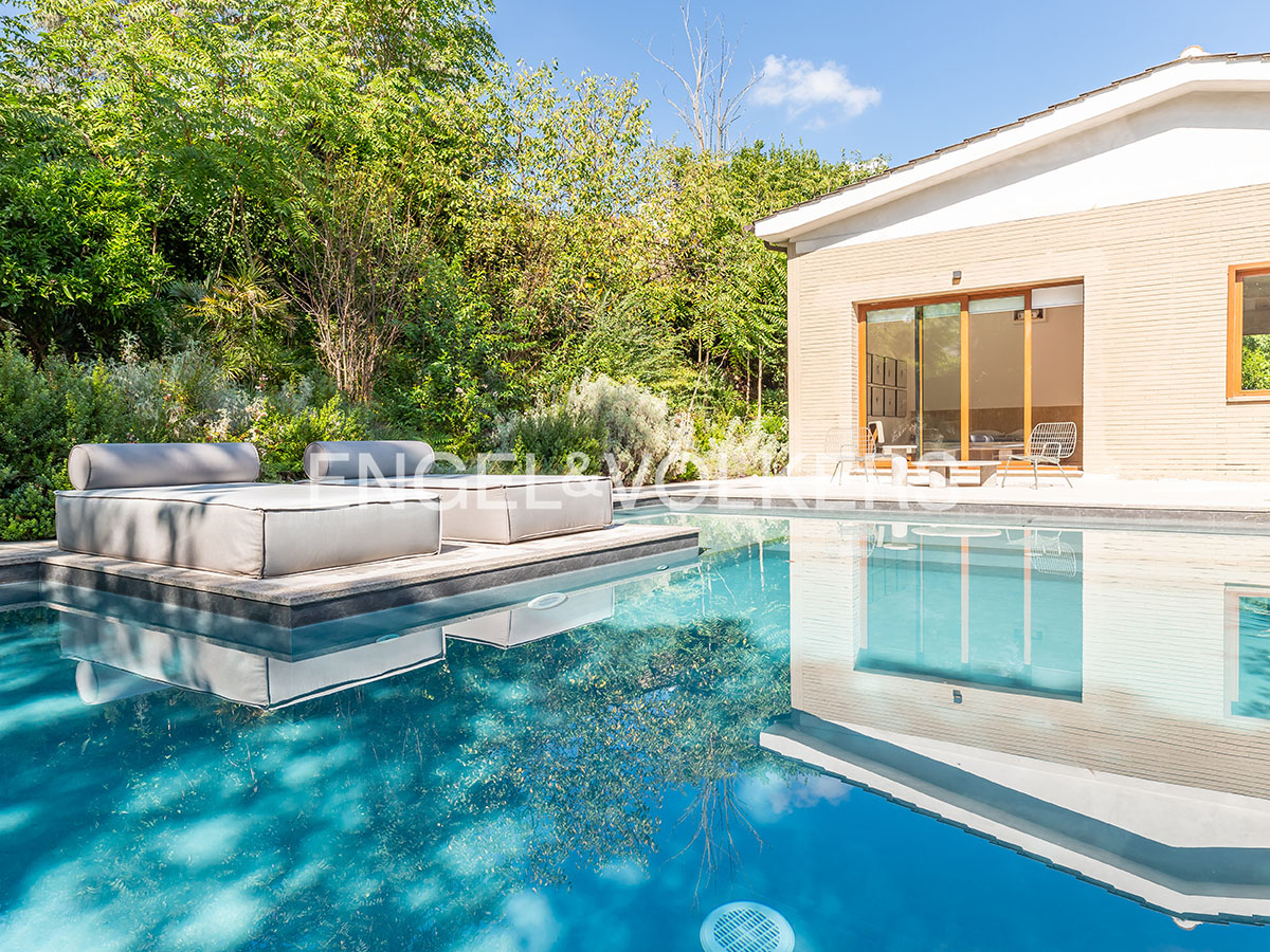 Outdoor pool with lounge chairs and a modern house in the background, reflecting in the water. Lush greenery surrounds the property.