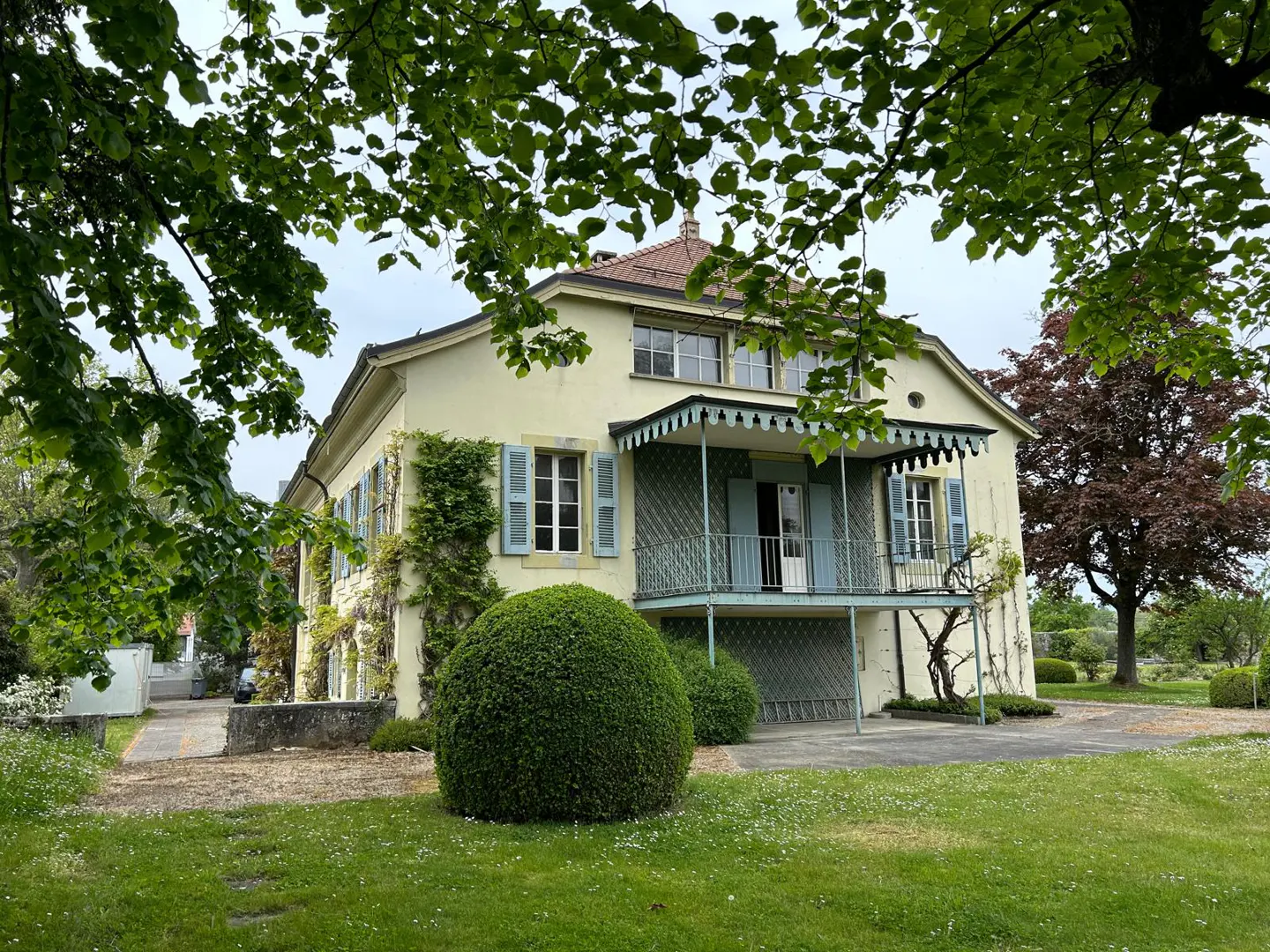 Two-story yellow house with blue shutters and a blue balcony, framed by green trees and lawn.