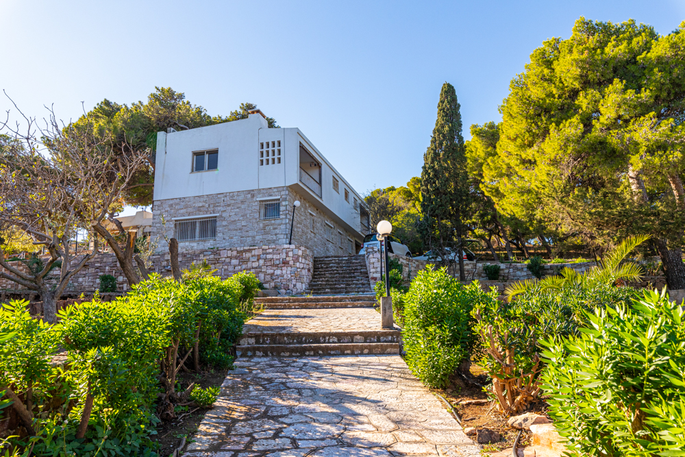 Exterior view of a two-story house with a stone path and stairs leading up to the entrance, surrounded by green trees and bushes.