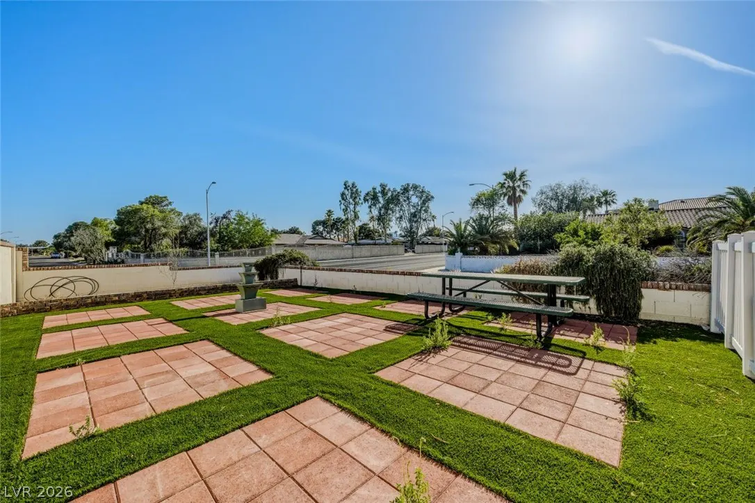 Outdoor patio with red pavers, green grass, and a picnic table under a clear blue sky. A tiered fountain sits in the grass.