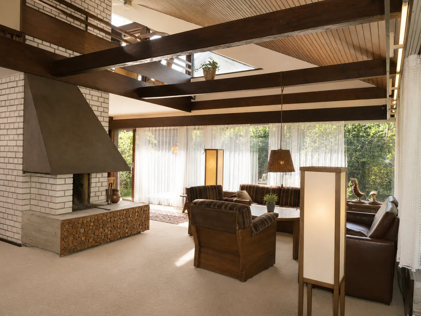 Living room with white brick fireplace, brown furniture, and wood-beamed ceiling. Large windows with sheer curtains let in natural light.