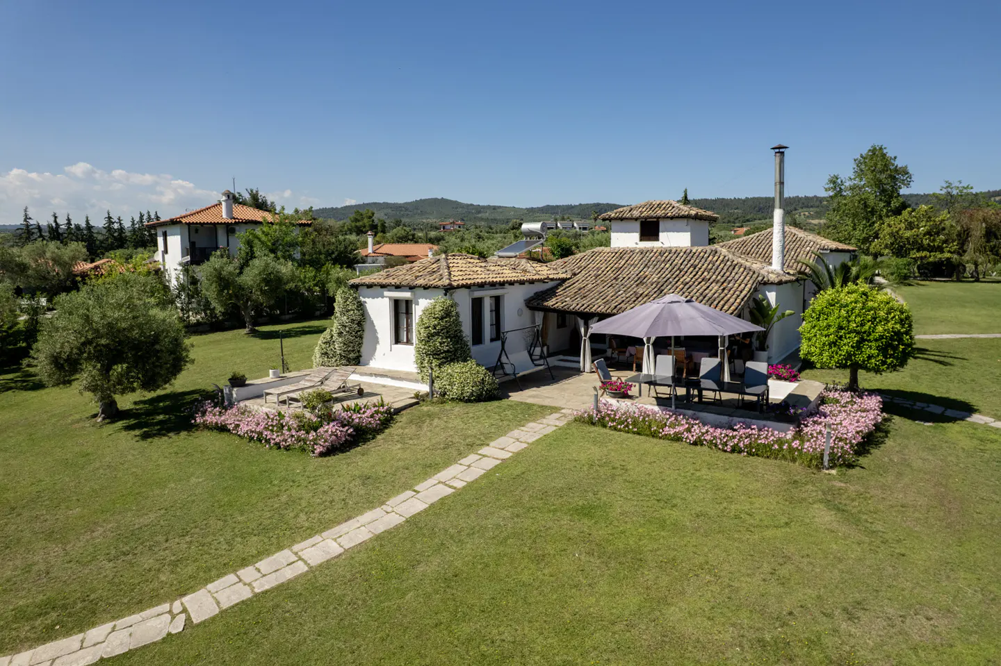 White Mediterranean villa with a tiled roof, patio, umbrella, and lush green lawn under a clear blue sky.