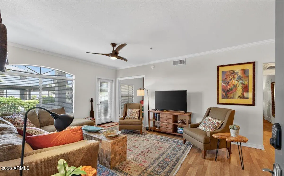 Living room with brown sofa, chairs, wood floors, and a patterned rug. A TV sits on a wooden console. Artwork hangs on the wall.