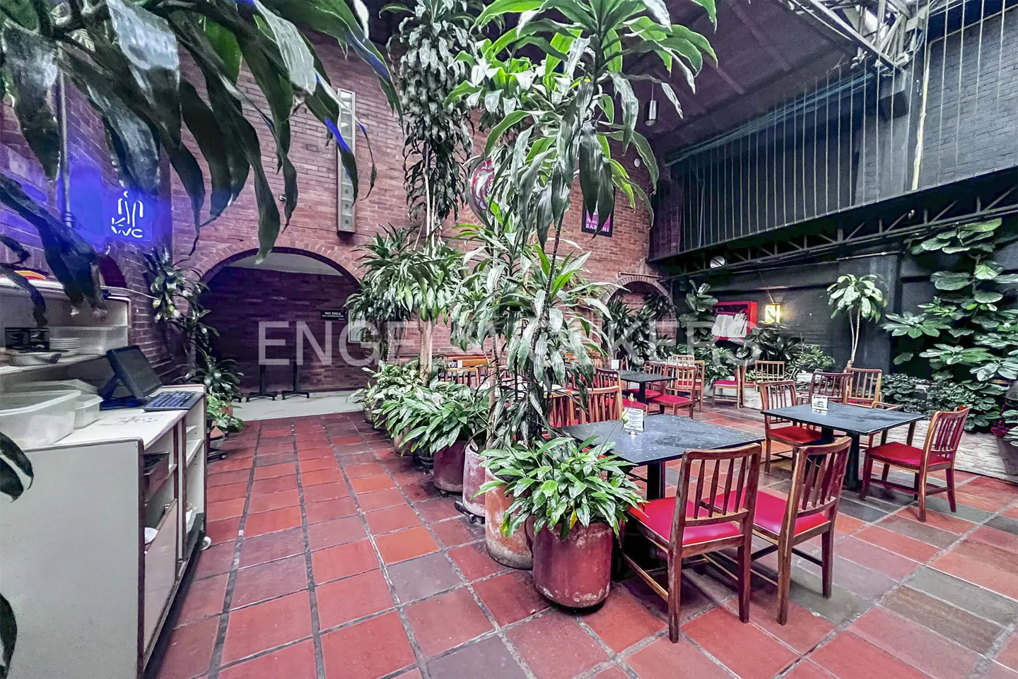 Indoor courtyard with red brick floor, tables, and chairs. Large potted plants and brick walls add to the ambiance.