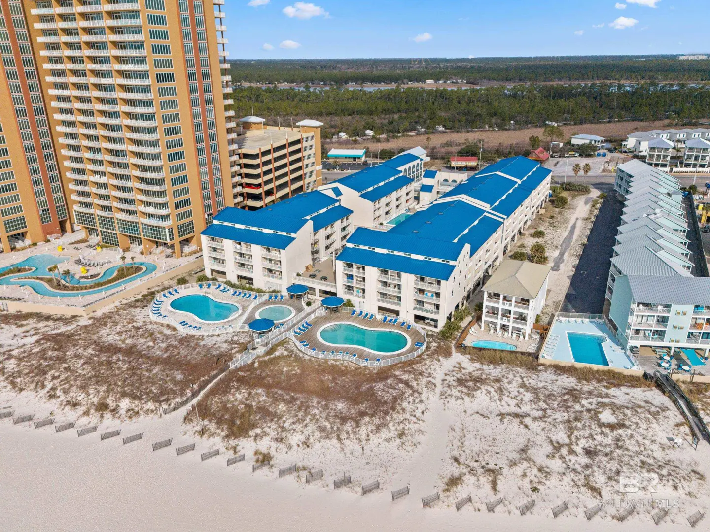 Aerial view of a beachside resort with white buildings, blue roofs, pools, and a tall orange condo tower.