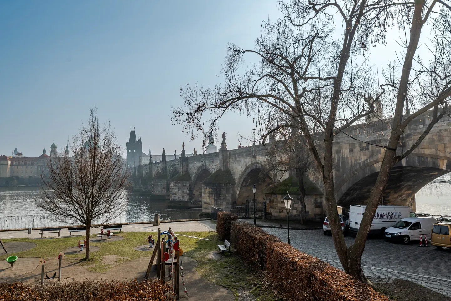 View of Prague's Charles Bridge from a park with playground equipment. Bare trees frame the stone bridge with statues. Cars are parked on the cobblestone street.