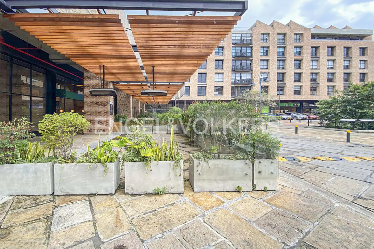 Exterior view of a brick building with a wooden awning and concrete planters. The ground is paved with stone.