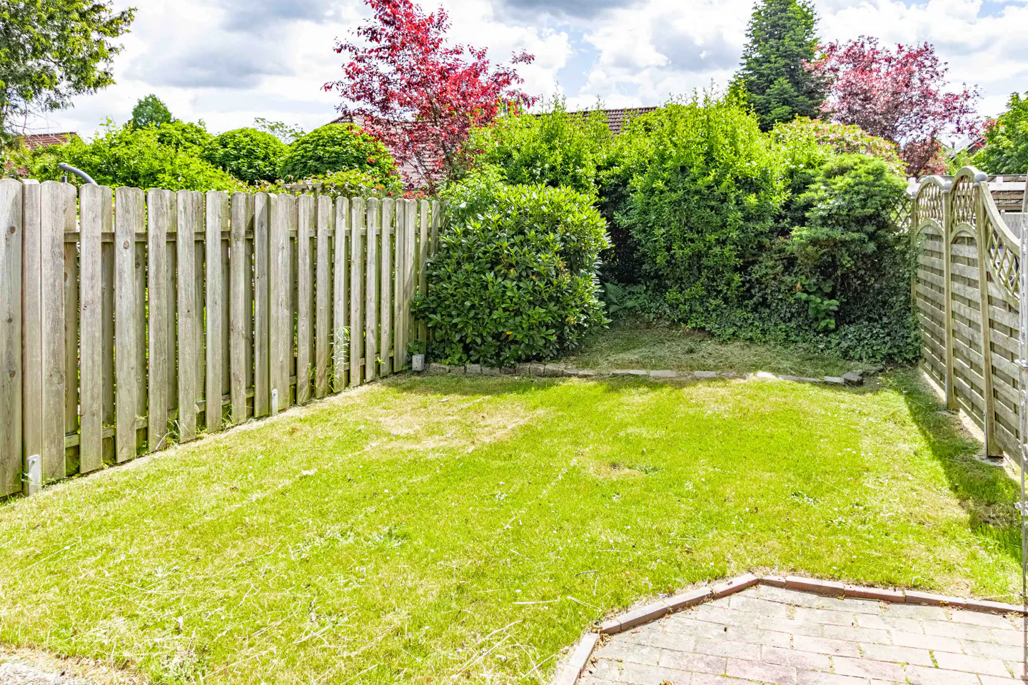 Small backyard with a green lawn, wooden fences on both sides, and lush green bushes under a cloudy sky.