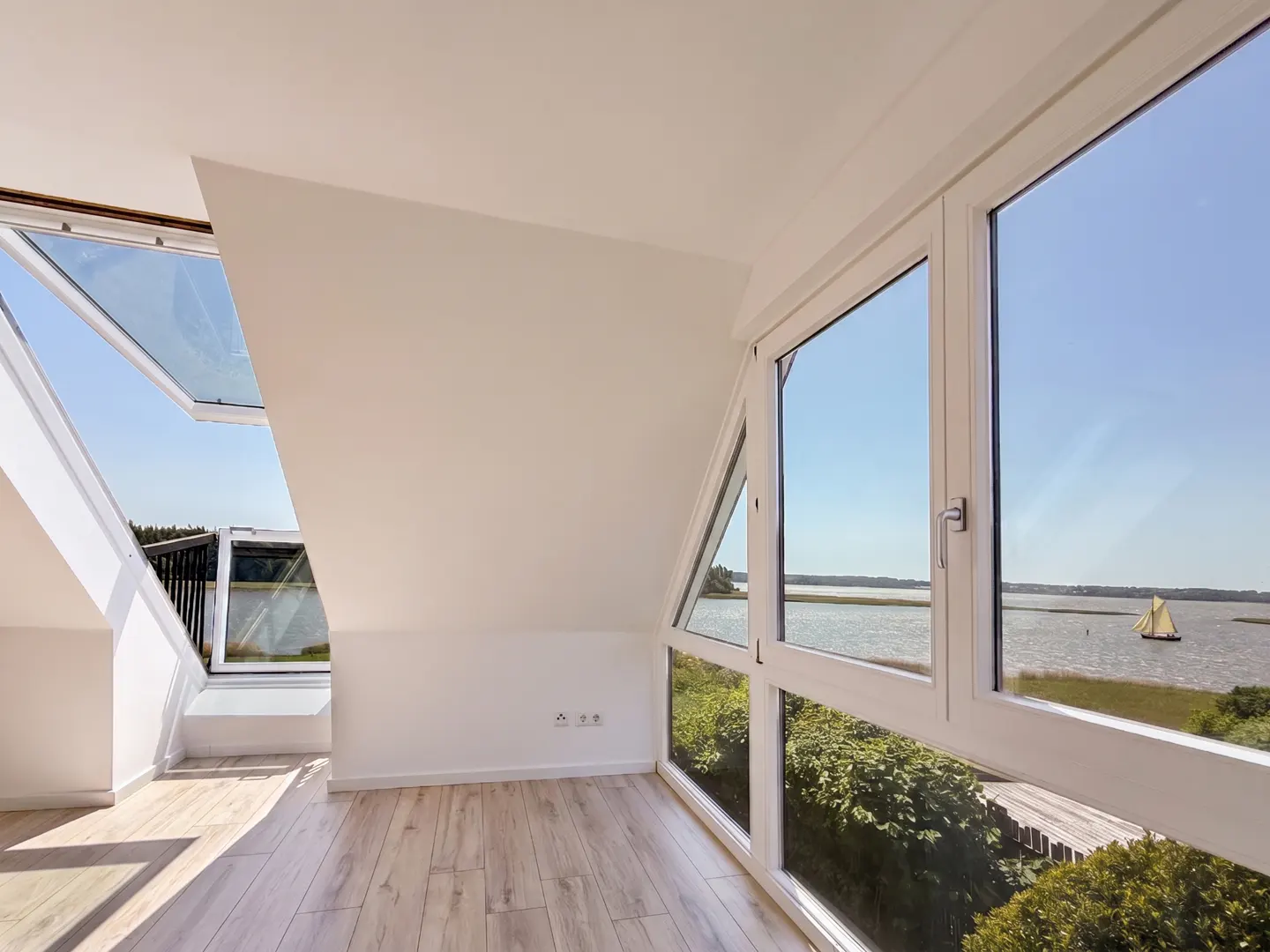 Bright attic room with white walls, wood floor, and large windows. View of water and sailboat. Skylight open to blue sky.