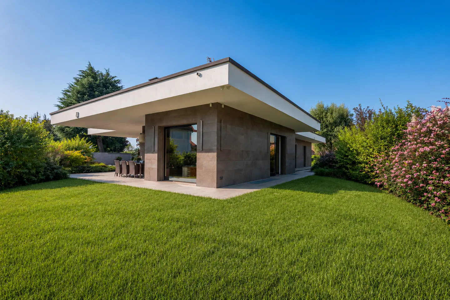 Modern single-story home with a flat roof, brown siding, and large windows, surrounded by green lawn and landscaping under a blue sky.