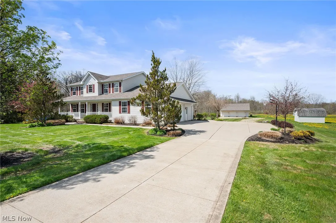 Two-story white house with red shutters, a long driveway, and green lawn under a blue sky.