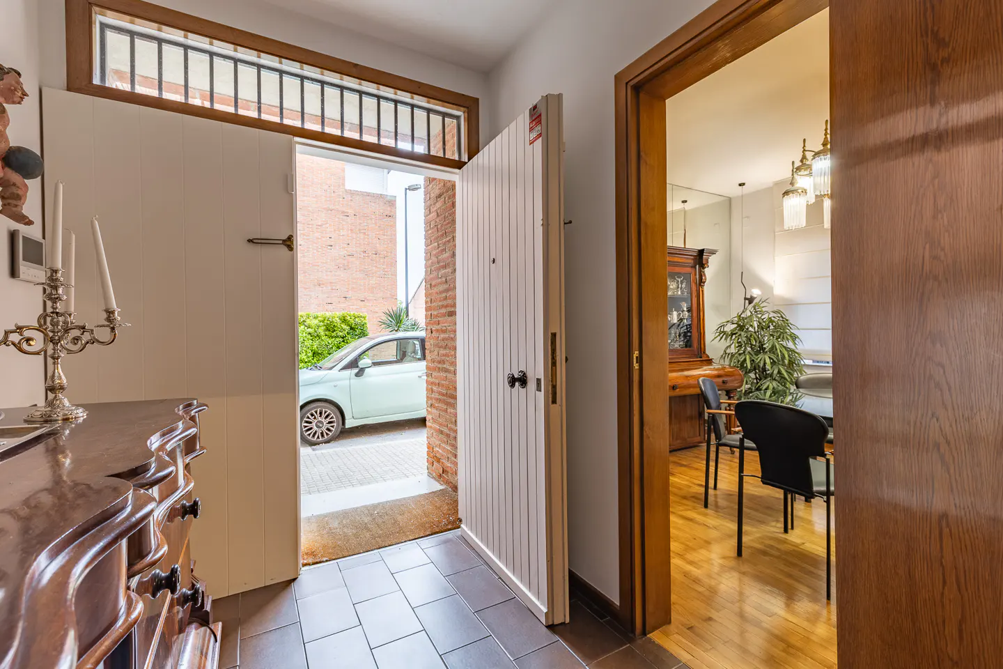Entryway with open white door, dark wood floors, and a glimpse of a light green car outside. A dining room is visible through another doorway.