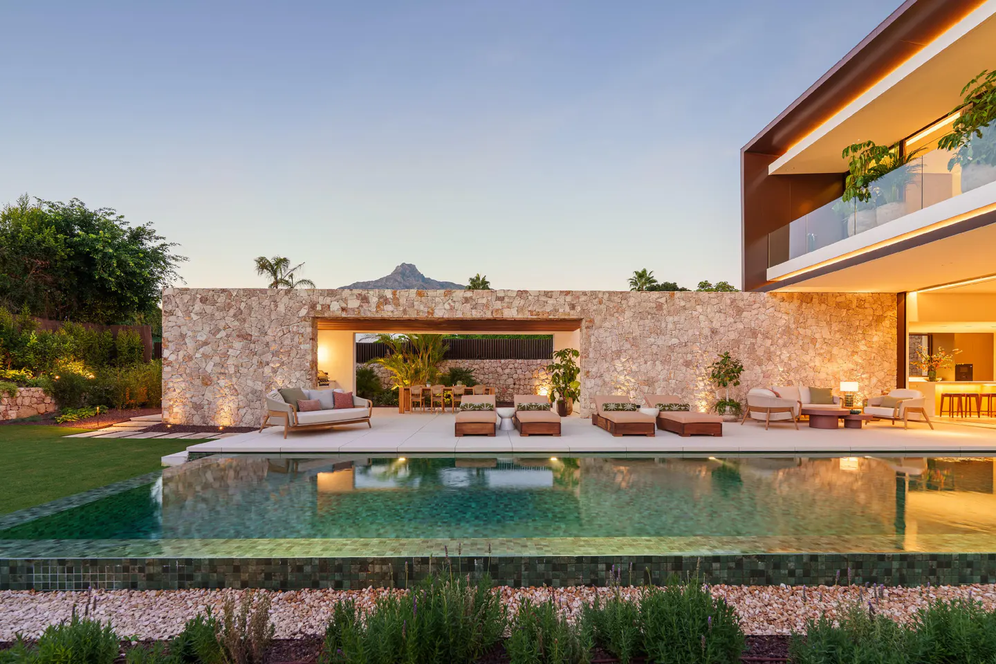 Luxury home exterior at dusk featuring a pool, stone wall, lounge furniture, and mountain backdrop.