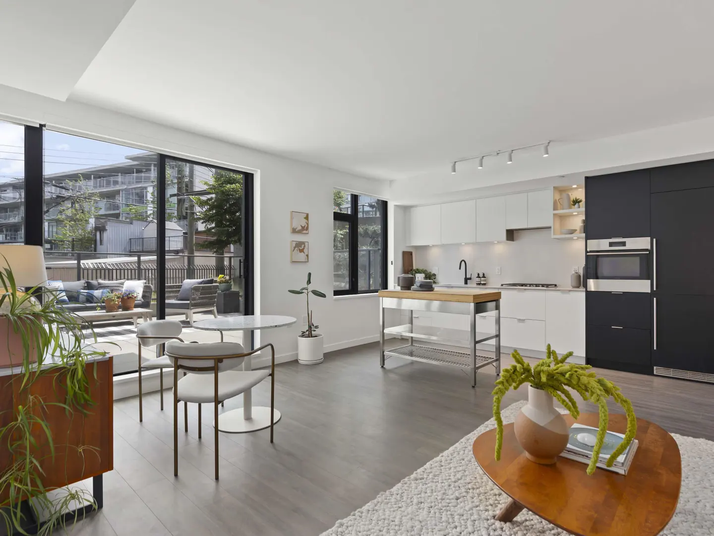 Bright, modern condo interior with white walls, gray floors, and black accents. Kitchen island, dining table, and balcony with outdoor seating.