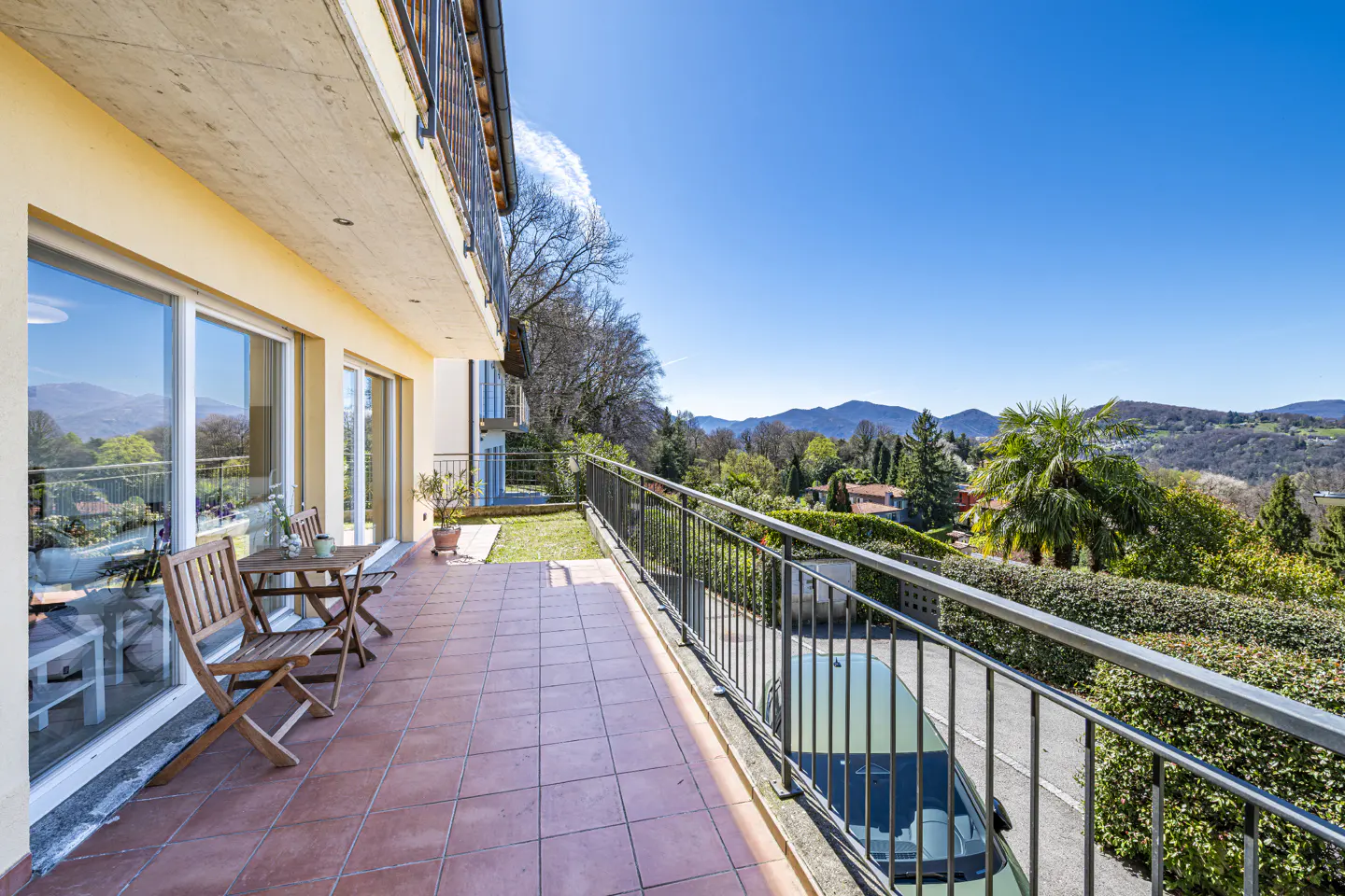 Balcony with terracotta tiles, metal railing, and wooden table set. Lush green landscape and blue sky in the background.