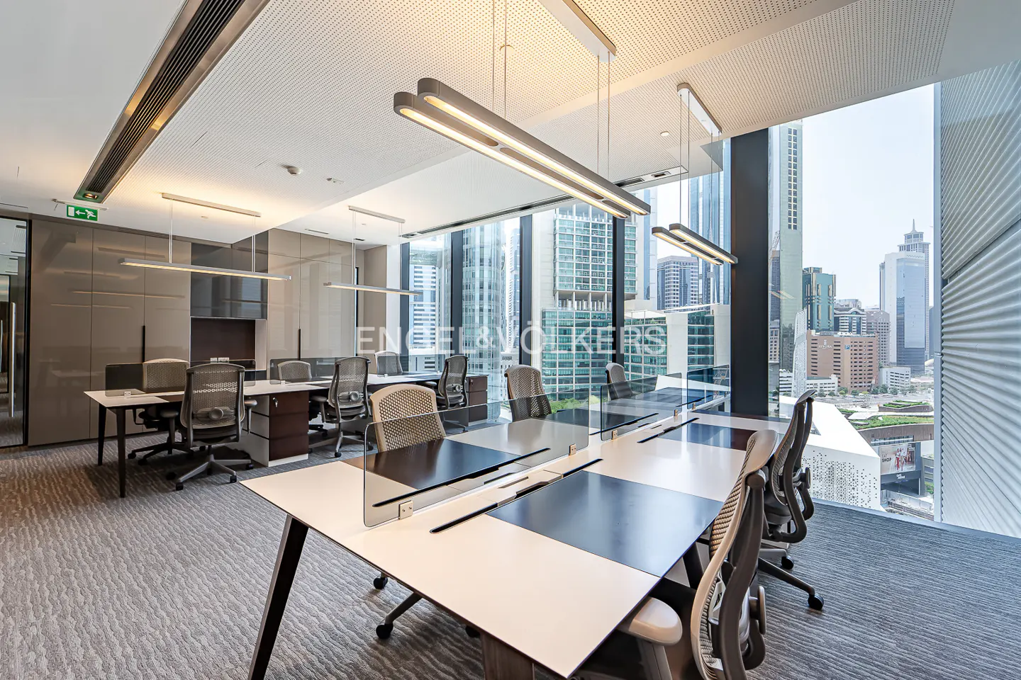 Bright, modern office space with desks, chairs, and large windows overlooking a city skyline. Linear lighting fixtures hang from the ceiling.
