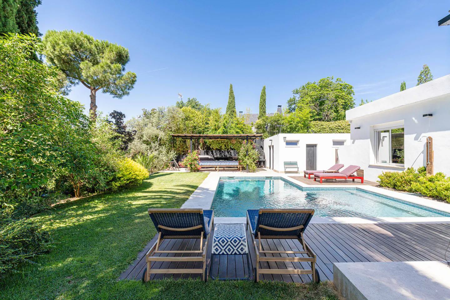 Backyard with a swimming pool, wooden deck, lounge chairs, and lush green garden under a clear blue sky.