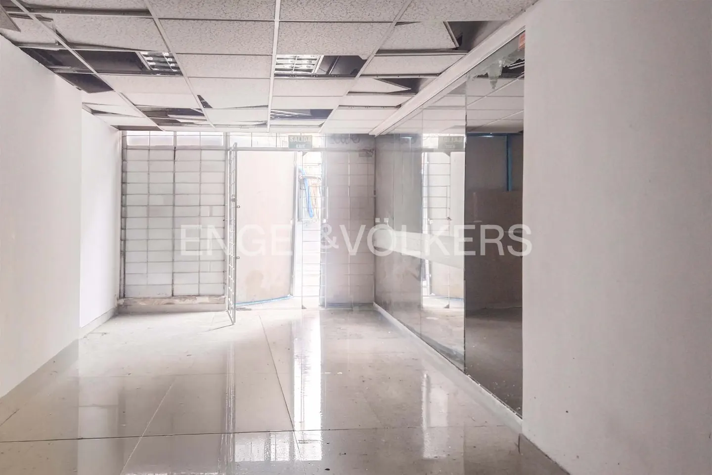 Interior view of a commercial space with white walls, a damaged ceiling, and a shiny floor. An open doorway leads to a brighter area. Engel & Völkers logo is visible.