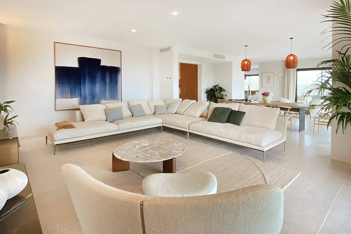 Bright, modern living room with a large white sectional sofa, marble coffee table, and blue abstract art. Dining area visible in background.
