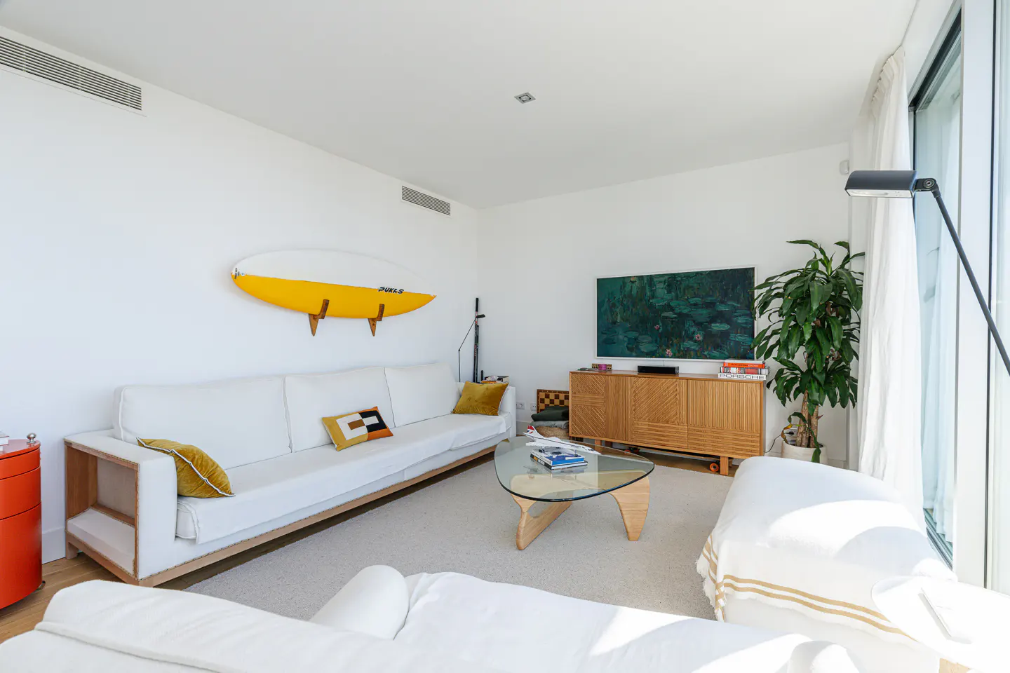 Bright living room with white sofa, glass coffee table, and yellow surfboard on the wall. A wooden cabinet and a large plant are near the window.