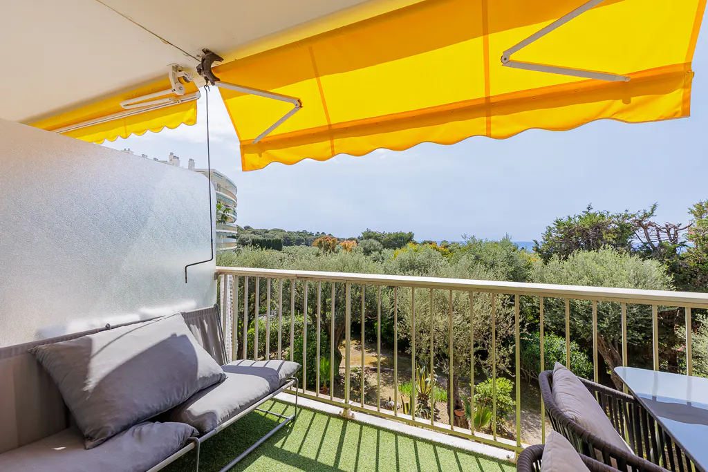Balcony view with yellow awning, gray cushions on a bench, and a metal railing overlooking lush greenery.