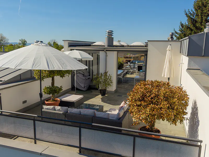 Rooftop patio with gray wicker furniture, white umbrellas, and potted plants. A glass door leads to an outdoor dining area.