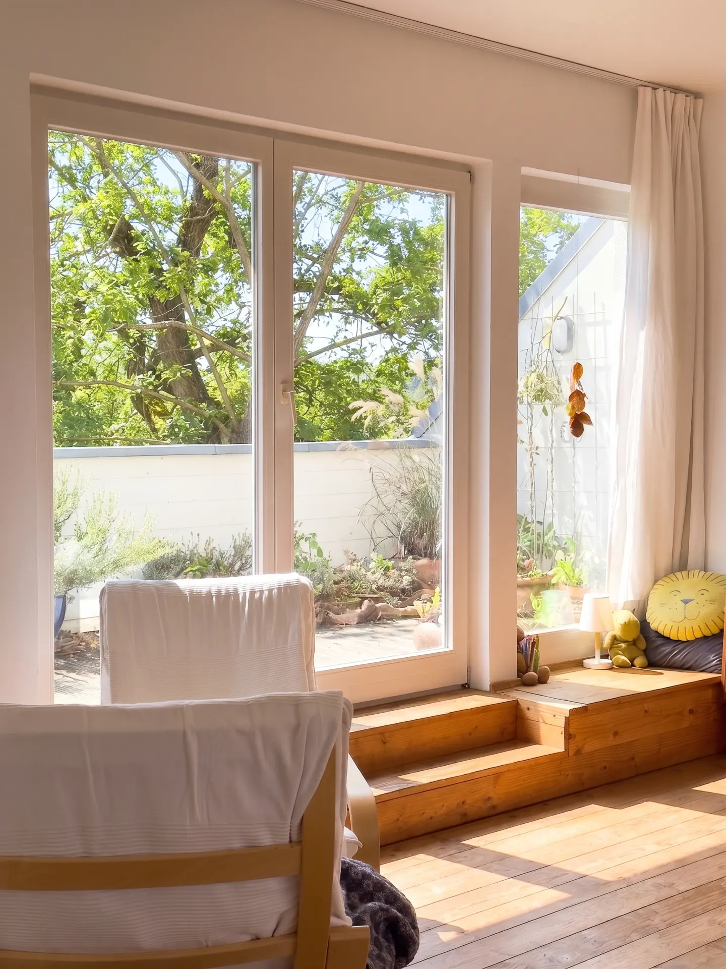 Bright room with wooden floors, a white sofa, and a large sliding glass door to a garden. A raised platform with toys sits by a window.