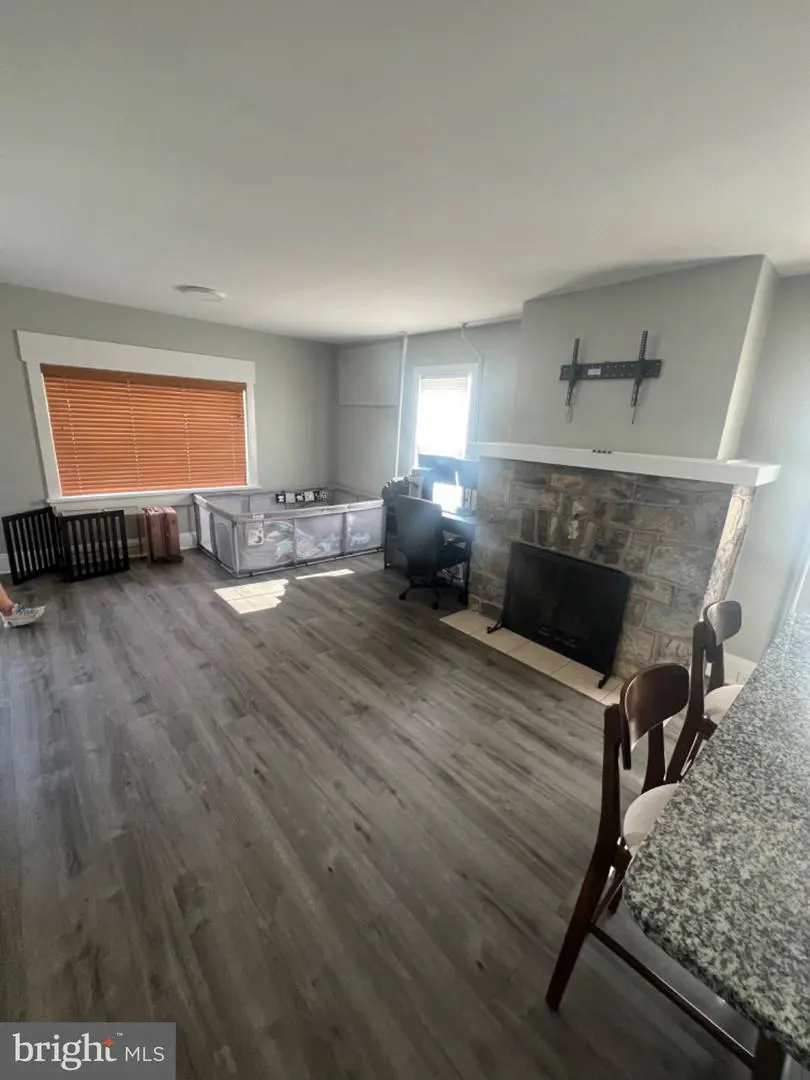 A living room with gray wood floors, a stone fireplace, and a playpen. A desk and chair are near the fireplace.