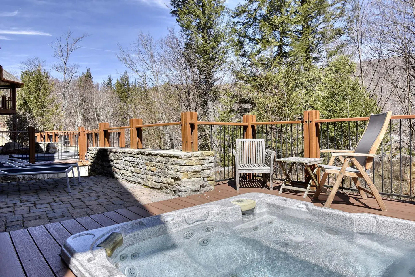 A hot tub on a wooden deck with patio chairs, a stone wall, and a scenic forest view under a clear blue sky.