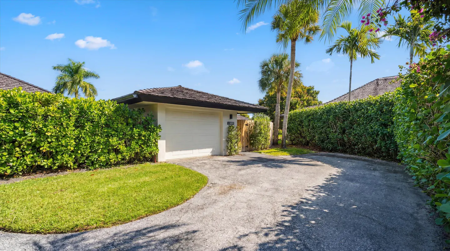 Exterior view of a white garage with a brown roof, surrounded by green hedges and palm trees under a blue sky.