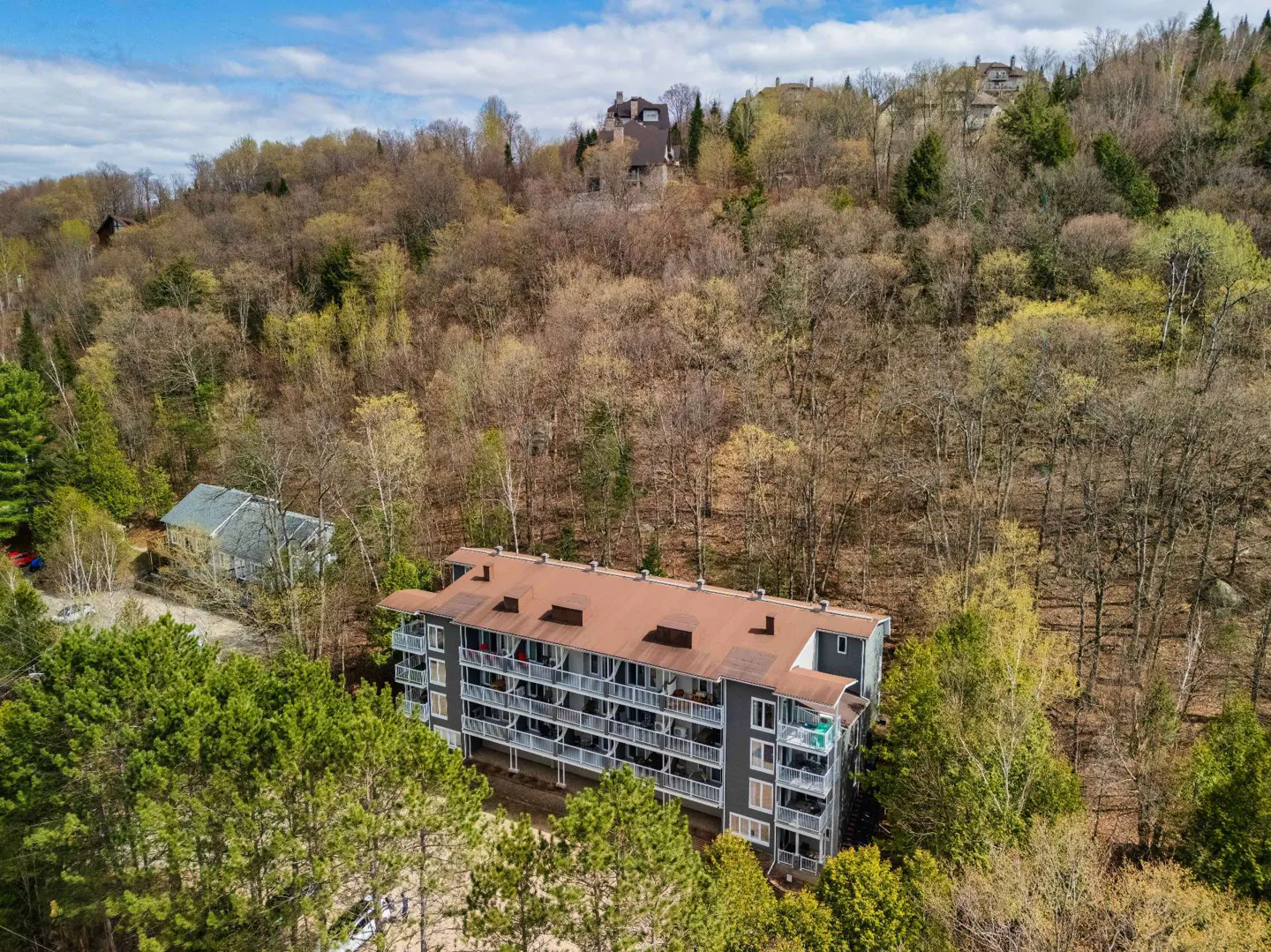 Aerial view of a gray condo building with brown roof, surrounded by green trees and a hillside with bare trees.