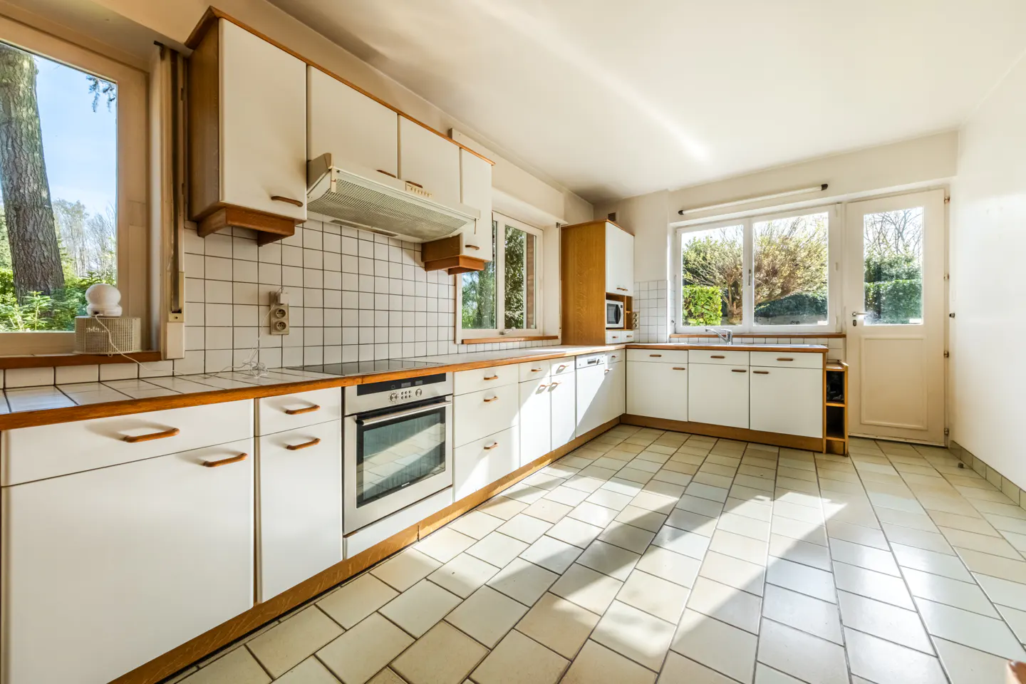 Bright kitchen with white cabinets, wood trim, and tile floors. Windows overlook a green yard. Oven and range are stainless steel.