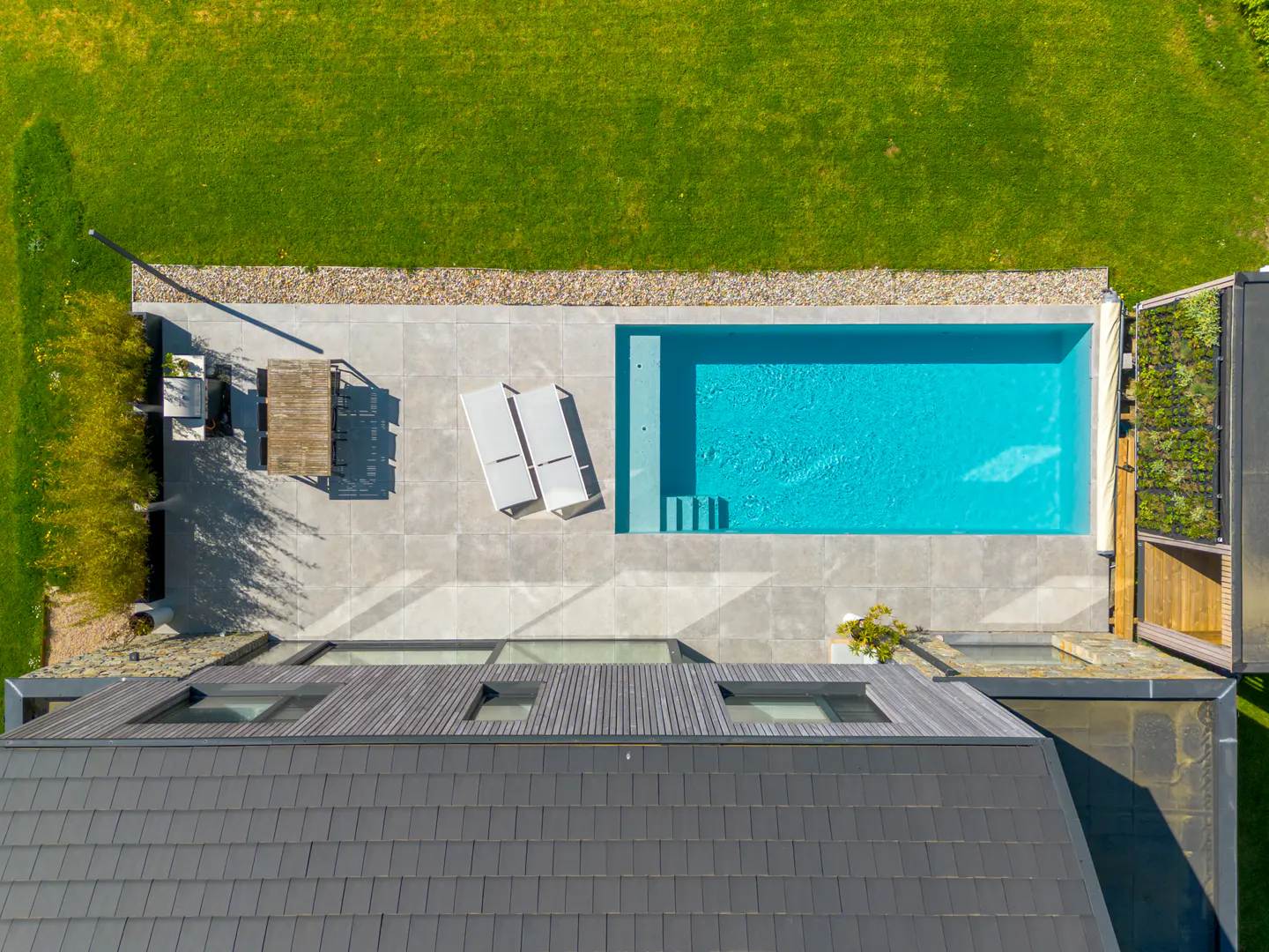Aerial view of a backyard with a blue swimming pool, gray patio, lawn, and a dark gray roof with skylights.