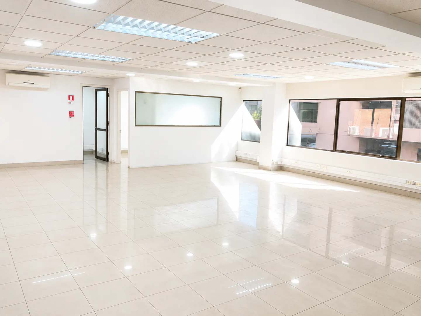 Bright, empty office space with white walls, glossy tiled floors, recessed ceiling lights, and large windows.