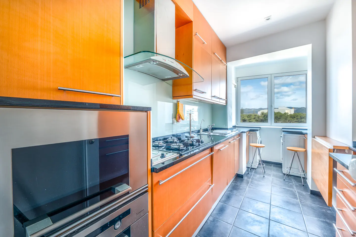 A modern kitchen with orange cabinets, black countertops, and stainless steel appliances. A window overlooks a green landscape.