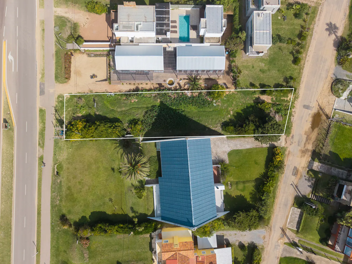 Aerial view of a rectangular vacant lot outlined in white, surrounded by green grass, trees, and houses.