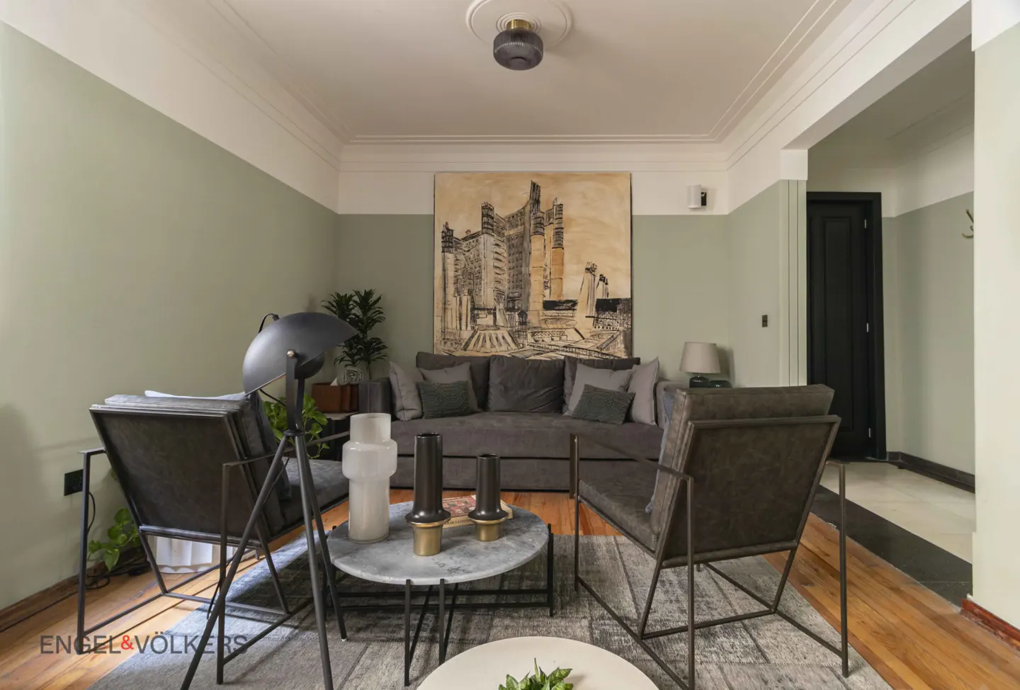 Living room with sage green walls, gray sofa, chairs, and a large building art piece. Two round tables sit on a gray rug.