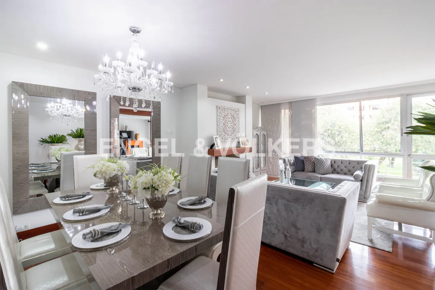 Open-concept living and dining area with wood floors, chandelier, and large windows. Dining table is set with white flowers and silver accents.