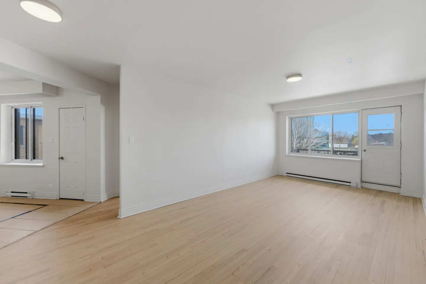 Bright, empty room with light wood floors, white walls, and two windows. One window has a door. A second room is visible to the left.
