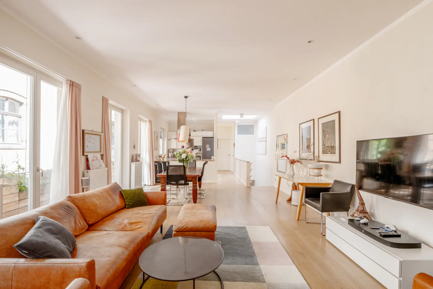 Bright open-plan living room with a tan leather sofa, round coffee table, dining area, and wall-mounted television.