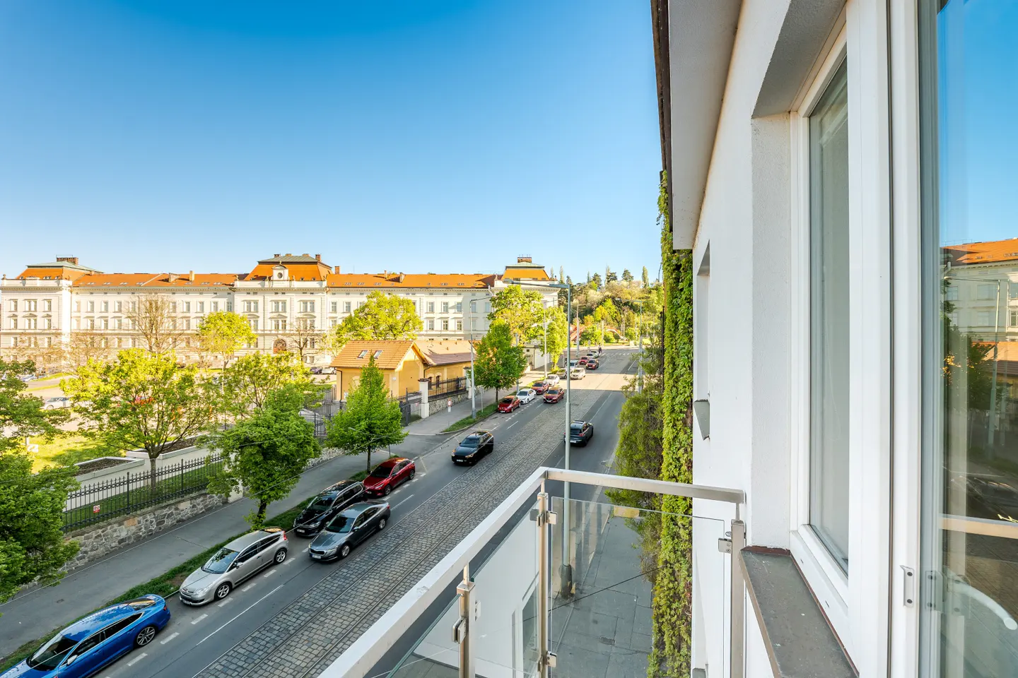 View from a balcony with a glass railing overlooking a street with cars, trees, and a large building in the background on a sunny day.