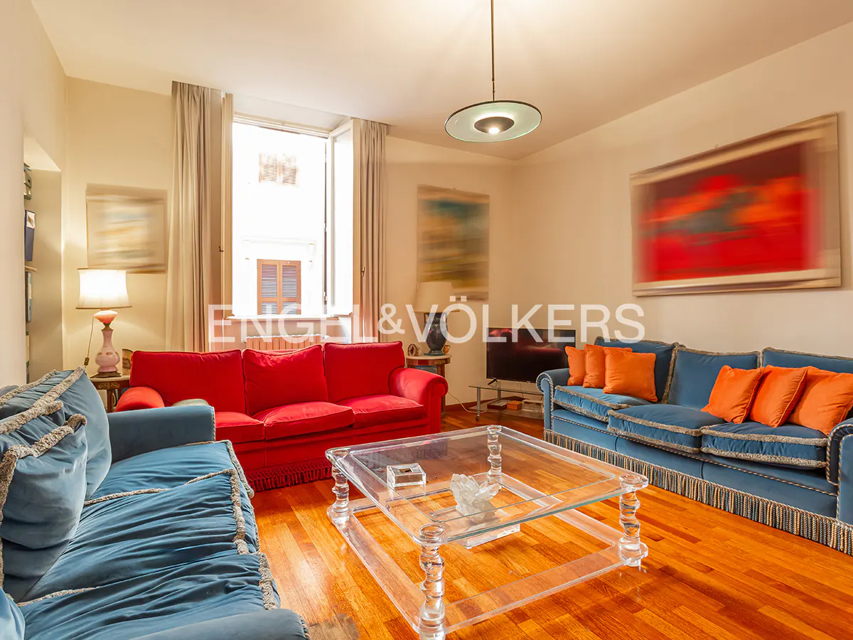 Bright living room with hardwood floors, blue and red sofas, orange pillows, and a clear acrylic coffee table.