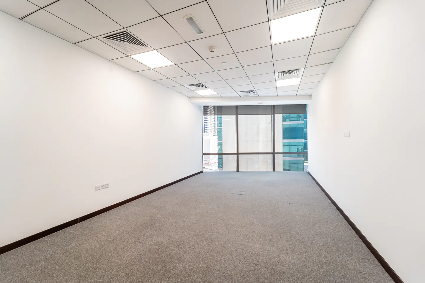 Empty office space with gray carpet, white walls, and a window view of city buildings.