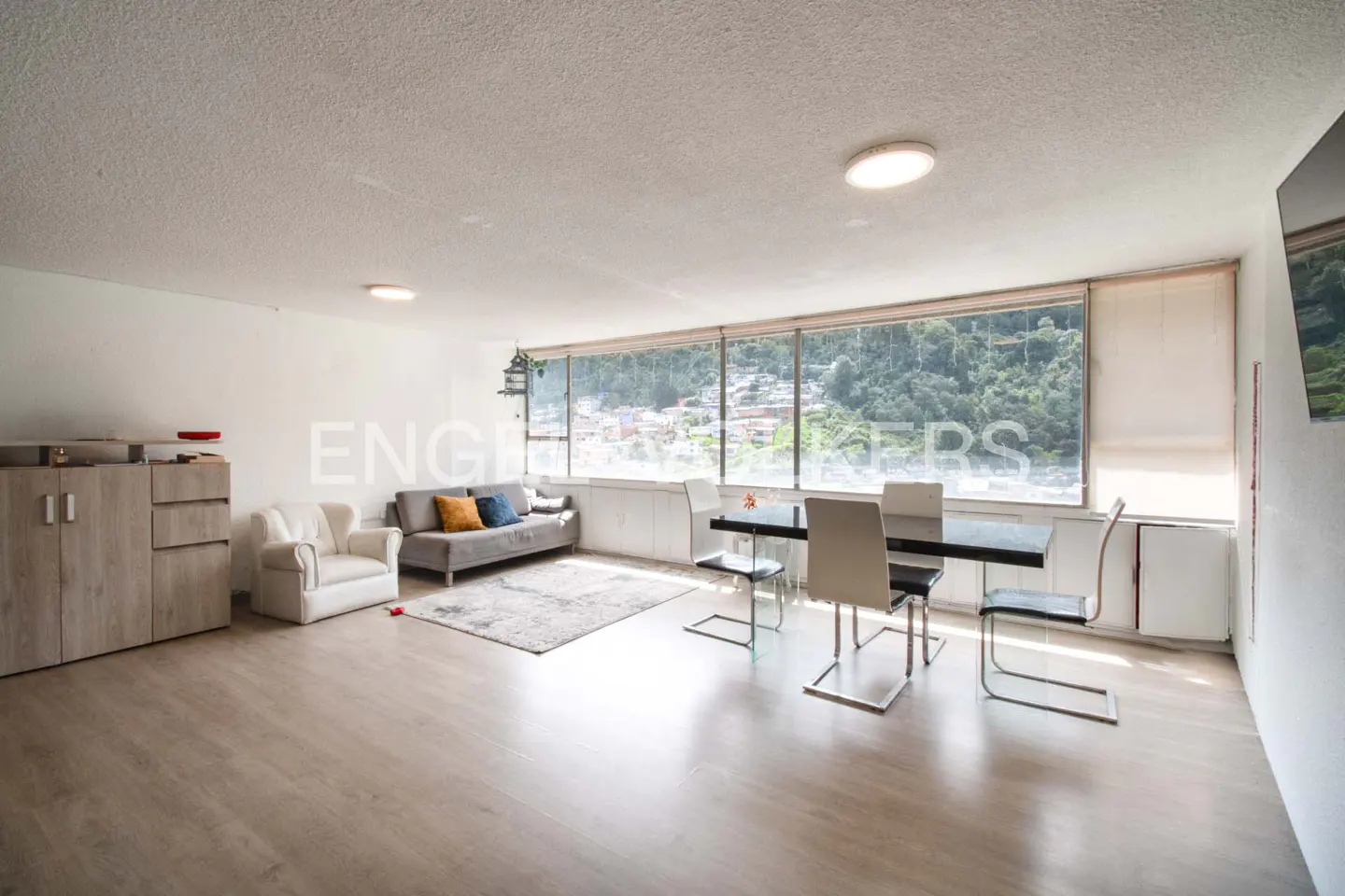 Bright, open-plan living and dining area with a grey sofa, white armchair, and a black dining table with white chairs. Large windows offer a view of a green, hilly landscape.