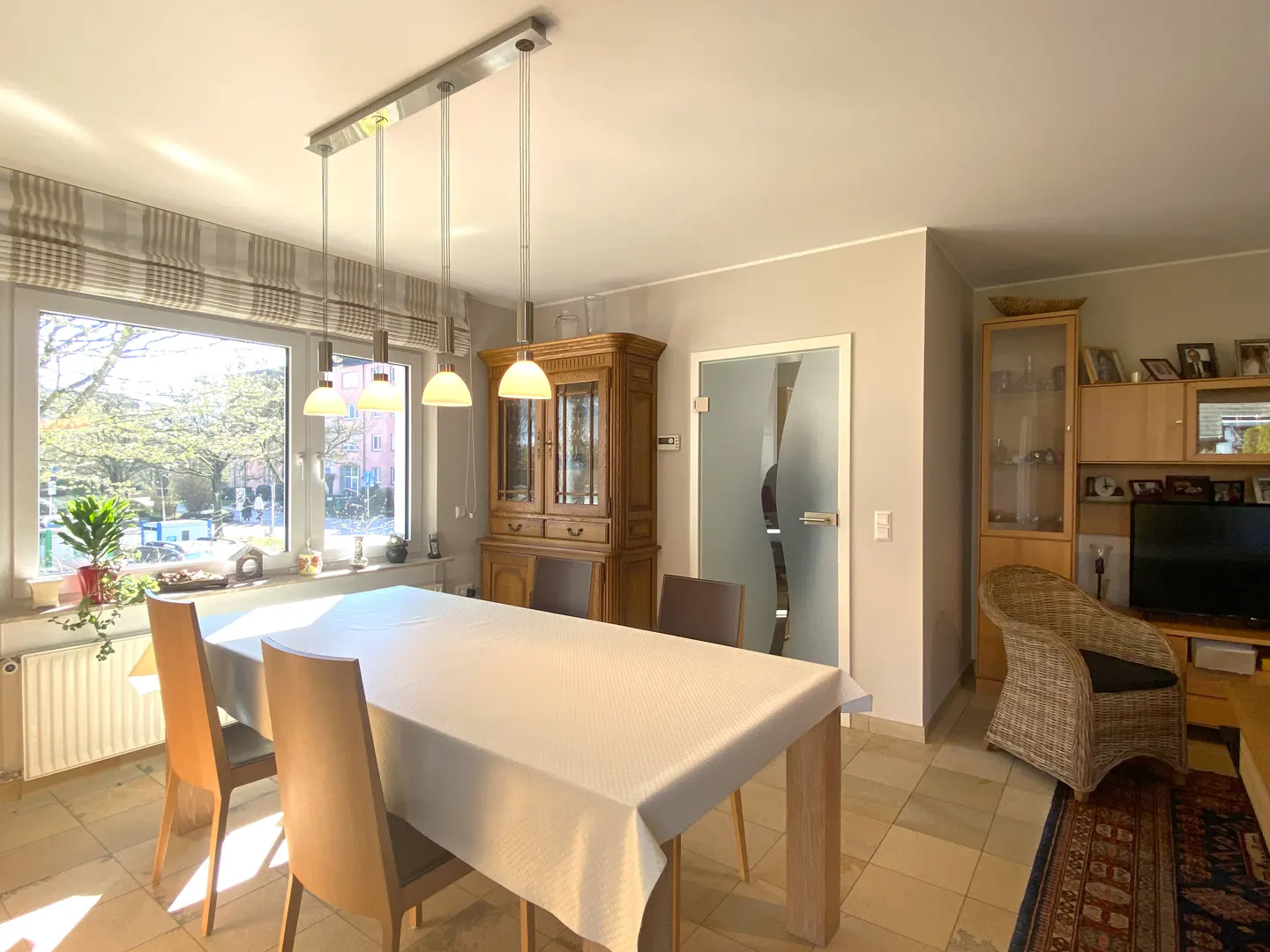 Bright dining room with a large window, a white tablecloth-covered table, and wooden chairs. A glass door and wooden cabinet are in the background.