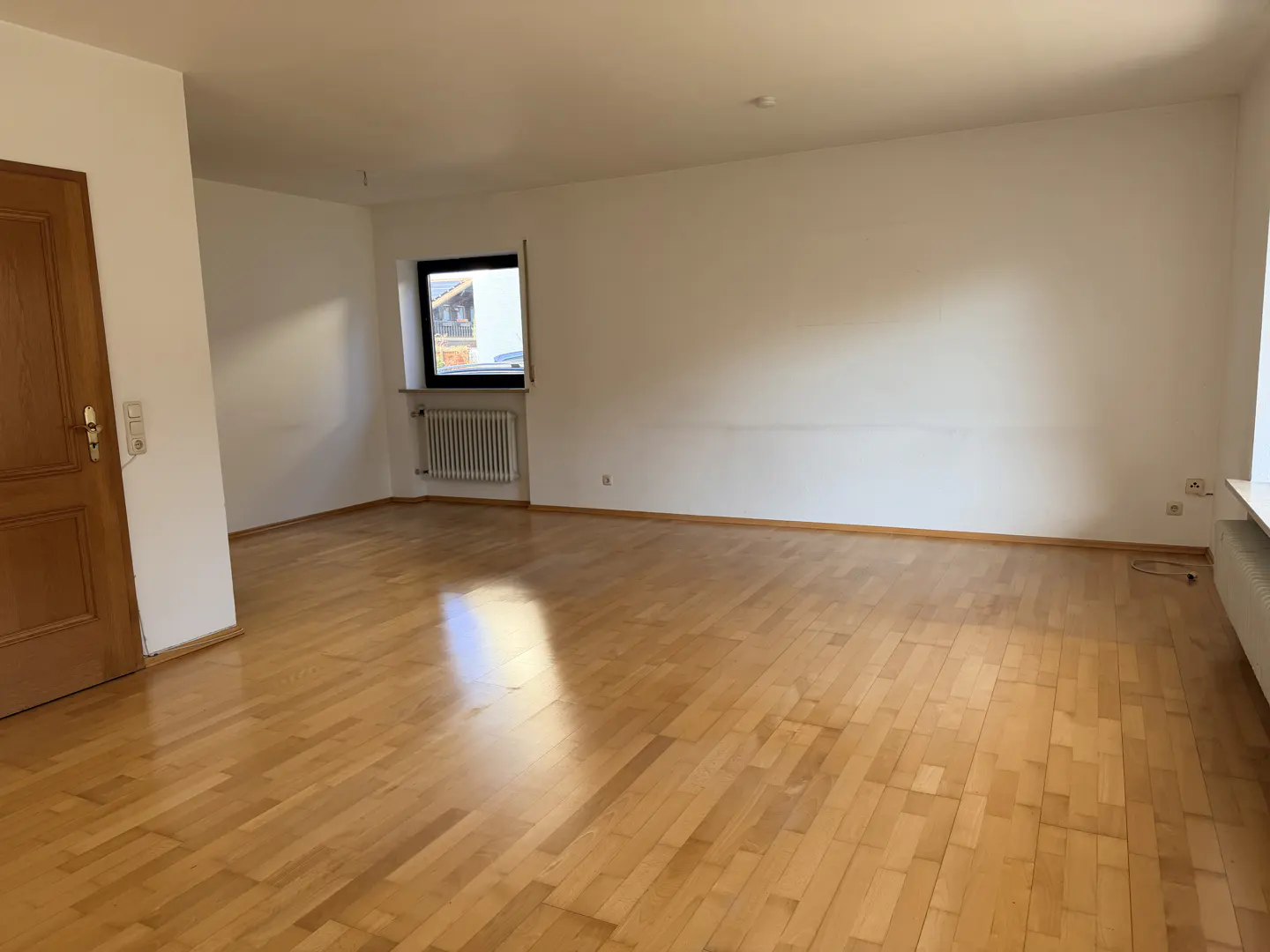 Empty room with light wood floors, white walls, and a wood door on the left. A window with a radiator is on the back wall.