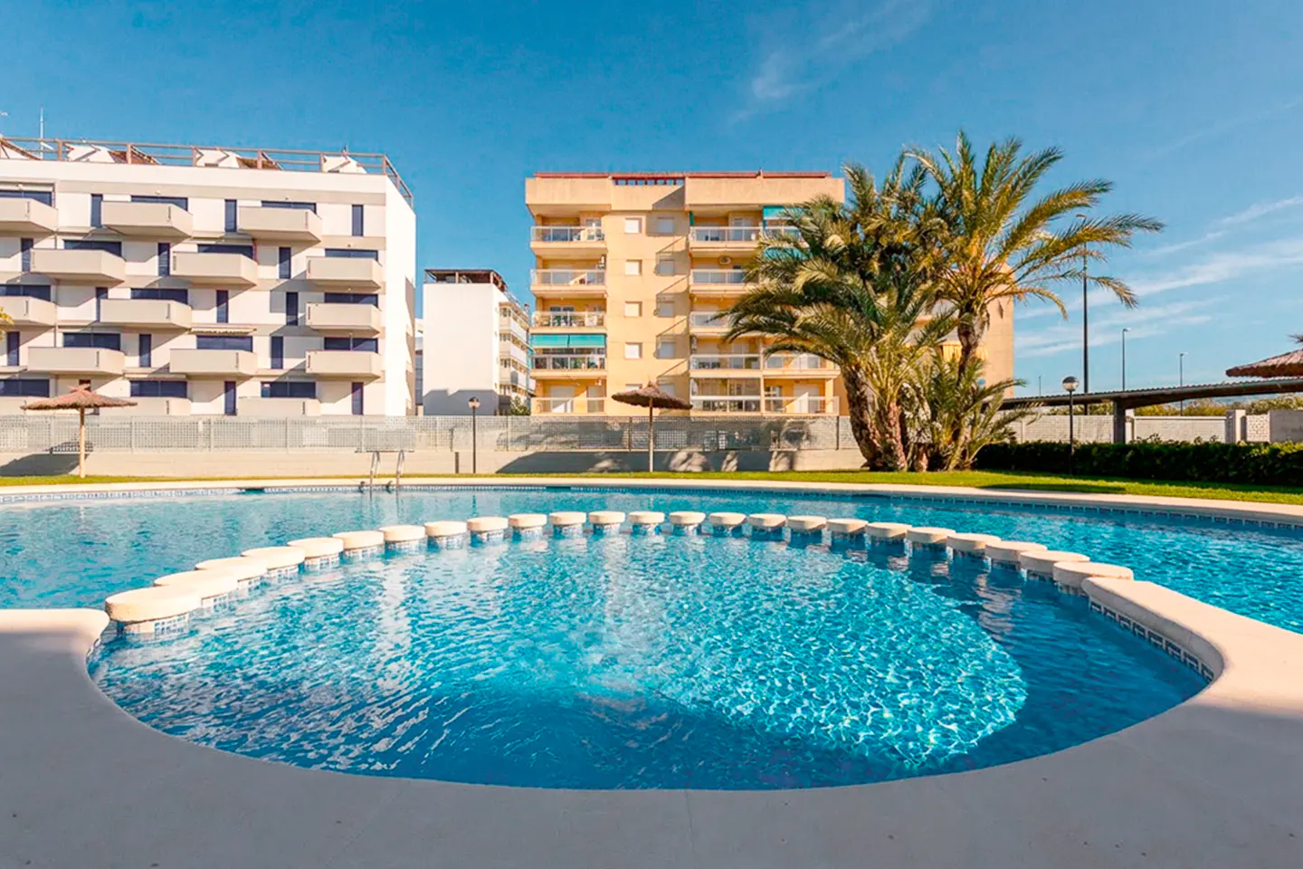 A circular swimming pool with blue water sits in front of modern apartment buildings and a palm tree under a clear blue sky.