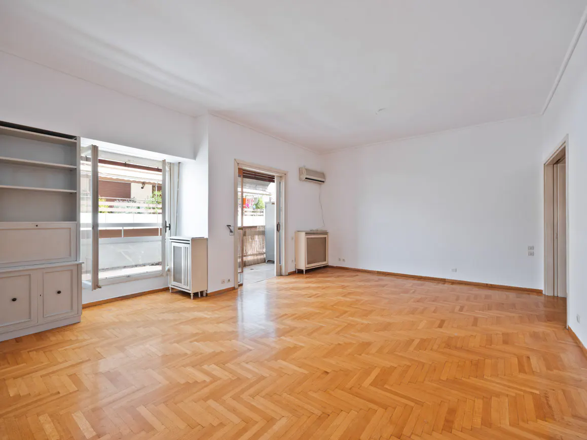 Bright, empty room with herringbone wood floors, white walls, and a balcony. A built-in bookcase sits to the left of the balcony doors.