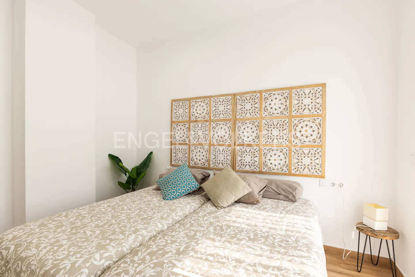 Bedroom with two twin beds, patterned headboard, blue and beige pillows, and a small wooden side table. A green plant sits in the corner.