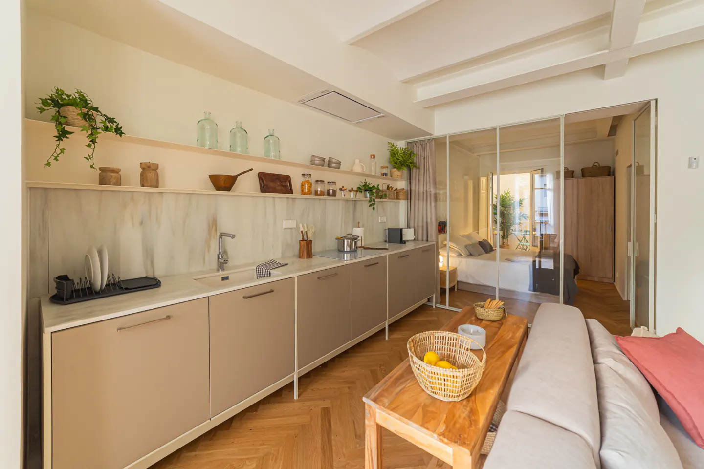Open-concept apartment with a kitchen featuring beige cabinets, open shelving, and a glass-enclosed bedroom in the background.